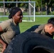 Two individuals in military-style olive/camouflage uniforms are performing a tire flip exercise on a grass field. A woman with braided hair and a black headband is visible on the left, while a man wearing glasses is pushing from the right side. The activity appears to be part of a physical training or fitness challenge event.