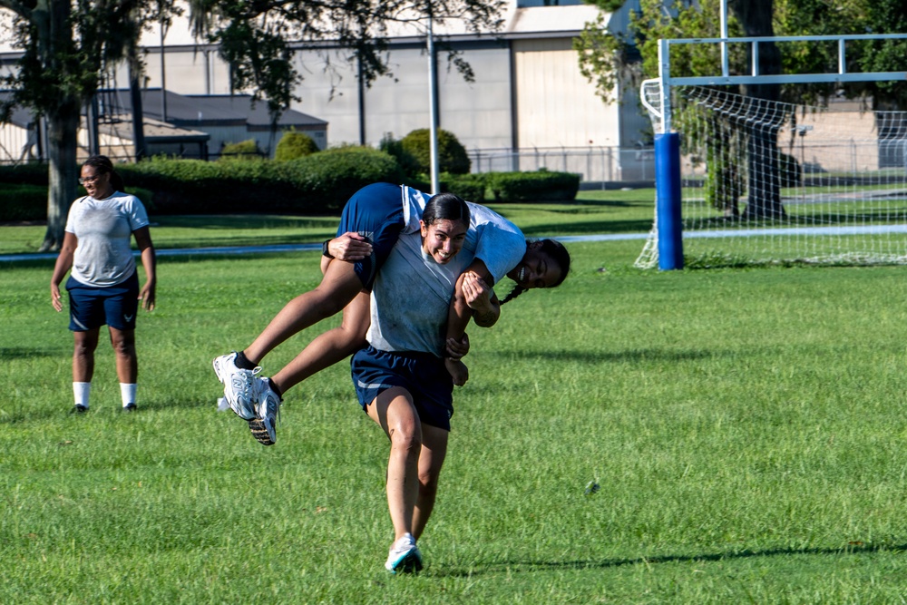 Three individuals are on a green grass athletic field, with one person carrying another on their back in what appears to be a partner carry exercise or drill. All individuals are wearing matching navy blue and gray athletic uniforms, suggesting a team or military/institutional fitness activity. A soccer goal post is visible in the background along with trees and a building.