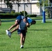 Three individuals are on a green grass athletic field, with one person carrying another on their back in what appears to be a partner carry exercise or drill. All individuals are wearing matching navy blue and gray athletic uniforms, suggesting a team or military/institutional fitness activity. A soccer goal post is visible in the background along with trees and a building.
