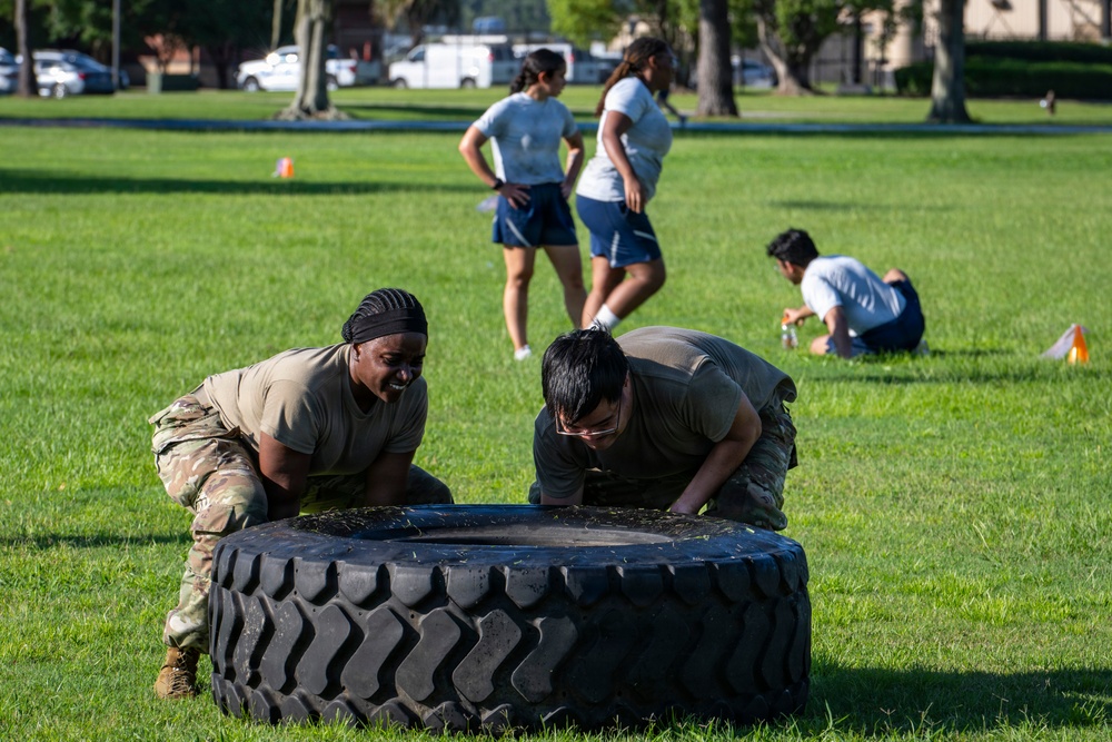Two individuals in military camouflage uniforms are performing a tire flip exercise on a grassy field, likely during a physical training session. In the background, several other participants in athletic wear are visible, along with orange marker cones indicating a structured fitness course or competition. The setting appears to be a military base or organized outdoor training area.