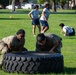 Two individuals in military camouflage uniforms are performing a tire flip exercise on a grassy field, likely during a physical training session. In the background, several other participants in athletic wear are visible, along with orange marker cones indicating a structured fitness course or competition. The setting appears to be a military base or organized outdoor training area.