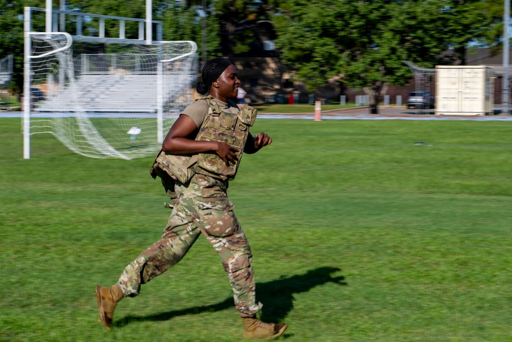 A military service member in full camouflage uniform and tactical vest is running across a grass field. The setting appears to be a military base or training facility, identifiable by the soccer/football goal post and institutional buildings in the background. The motion blur on the background suggests the subject is moving at a significant pace.