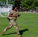 A military service member in full camouflage uniform and tactical vest is running across a grass field. The setting appears to be a military base or training facility, identifiable by the soccer/football goal post and institutional buildings in the background. The motion blur on the background suggests the subject is moving at a significant pace.
