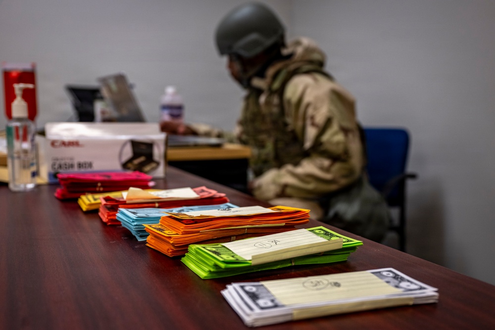A person wearing military/tactical gear including a helmet, body armor, and camouflage uniform is seated at a desk working on a computer or paperwork. In the foreground, multiple stacks of colorful envelopes or cards are arranged in rows on a wooden table. A cash box and hand sanitizer are also visible on the desk in the background.