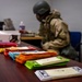 A person wearing military/tactical gear including a helmet, body armor, and camouflage uniform is seated at a desk working on a computer or paperwork. In the foreground, multiple stacks of colorful envelopes or cards are arranged in rows on a wooden table. A cash box and hand sanitizer are also visible on the desk in the background.