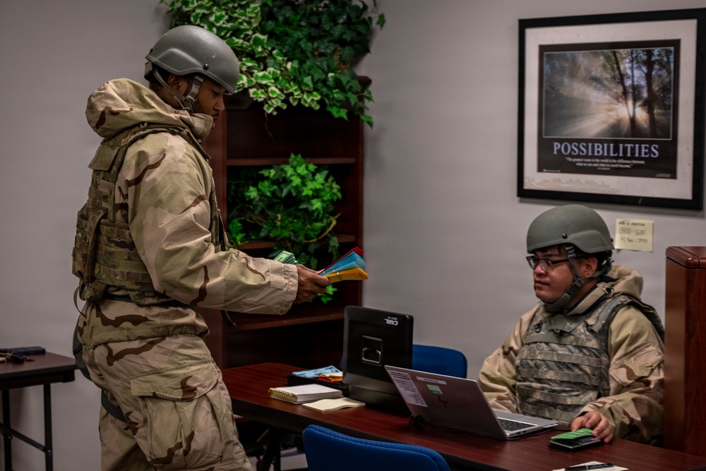 Two individuals in military combat uniforms and helmets are engaged in what appears to be a training exercise or operational briefing inside an office environment. The standing person is reviewing colorful cards or documents while the seated person works at a laptop computer. The setting combines a standard office interior with military gear, suggesting a simulated or real-world operational scenario.
