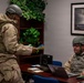 Two individuals in military combat uniforms and helmets are engaged in what appears to be a training exercise or operational briefing inside an office environment. The standing person is reviewing colorful cards or documents while the seated person works at a laptop computer. The setting combines a standard office interior with military gear, suggesting a simulated or real-world operational scenario.