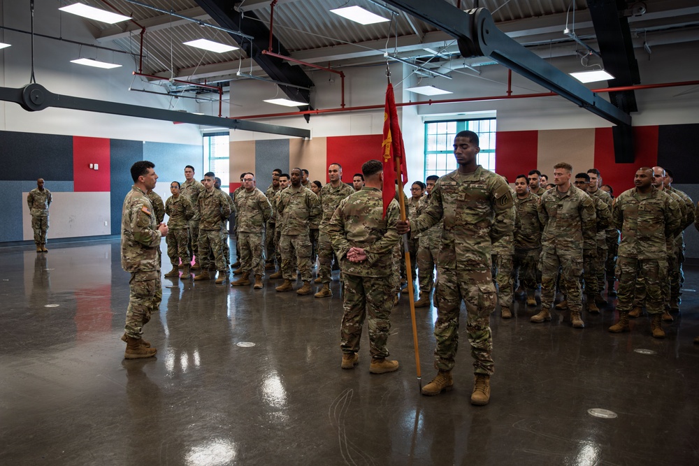 A formation of U.S. Army soldiers in OCP (Operational Camouflage Pattern) uniforms stands at attention inside a modern indoor facility. A soldier in the foreground holds a red unit guidon on a wooden staff, while an officer or NCO addresses the formation from the left. The setting appears to be a formal military ceremony or briefing, likely a unit formation, activation, or change of command event.