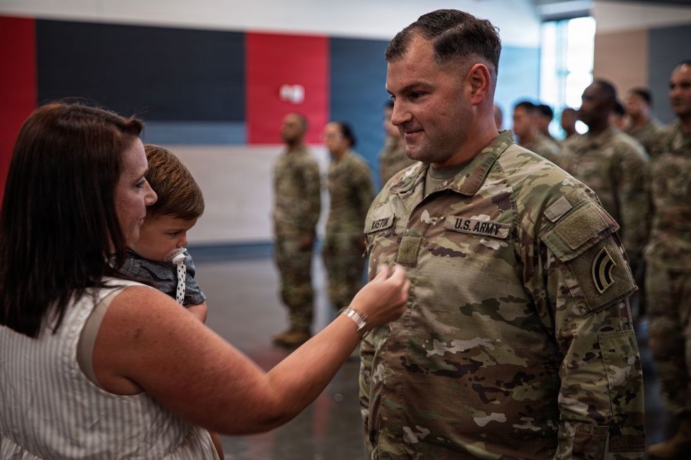 A U.S. Army soldier is having an insignia or pin affixed to his uniform by a woman holding a young child, likely during a promotion or award ceremony. The soldier's name tape reads 'KASTON' and he wears Staff Sergeant rank insignia on his right sleeve. Other uniformed soldiers are visible standing in formation in the background.