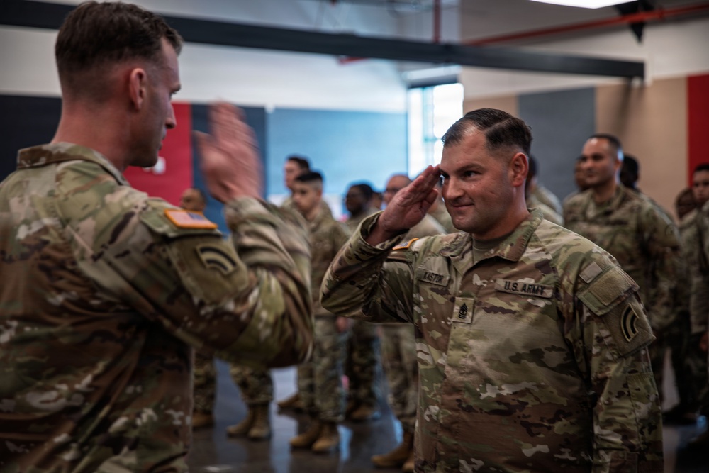 A U.S. Army ceremony is taking place indoors, likely a promotion or award ceremony. A soldier identified by nametape as 'LISTIN' with sergeant first class rank insignia is rendering a salute to an officer facing away from the camera. Multiple soldiers in OCP (Operational Camouflage Pattern) uniforms are standing in formation in the background.