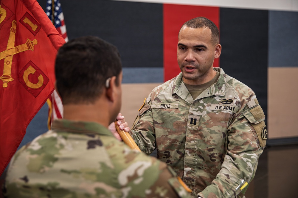 A U.S. Army Captain named Ortiz is participating in what appears to be a military change of command or guidon ceremony, receiving or passing a unit guidon staff to/from another soldier. Both individuals are wearing OCP (Operational Camouflage Pattern) uniforms. A red Artillery unit guidon and an American flag are visible in the background.