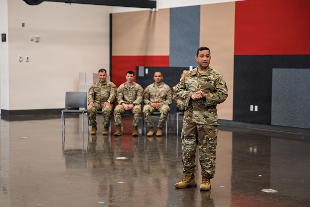 A U.S. Army soldier, identified by a name tape reading 'DELGADO,' stands in the foreground addressing or briefing a group of four fellow soldiers seated in chairs behind him. The setting appears to be a large indoor facility with polished concrete floors and acoustic panel-covered walls in red, tan, and gray. All personnel are wearing OCP (Operational Camouflage Pattern) uniforms.