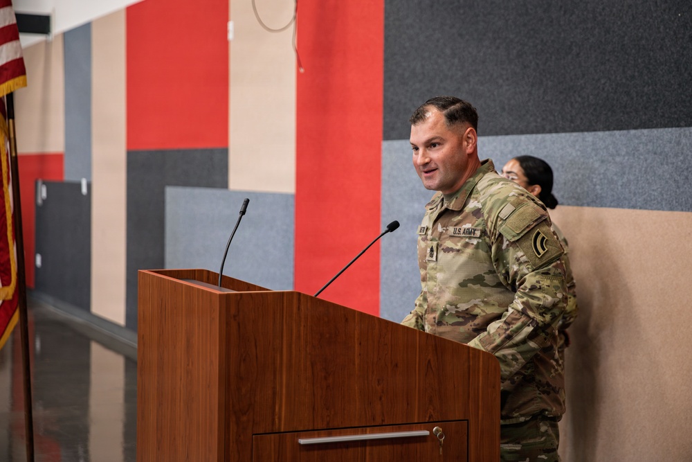 A U.S. Army soldier in Operational Camouflage Pattern (OCP) uniform is speaking at a wooden podium equipped with two microphones. The setting appears to be an indoor military ceremony or briefing room with colorful acoustic panels on the wall and an American flag visible to the left. A second uniformed individual is partially visible in the background.