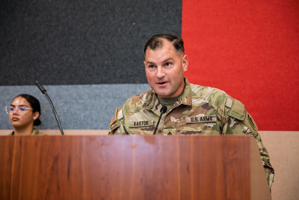 A U.S. Army soldier named Kastor is speaking at a wooden podium with a microphone. He is wearing an OCP (Operational Camouflage Pattern) uniform with 'U.S. ARMY' and 'KASTOR' name tapes visible. A second person in military uniform is seated in the background to the left.