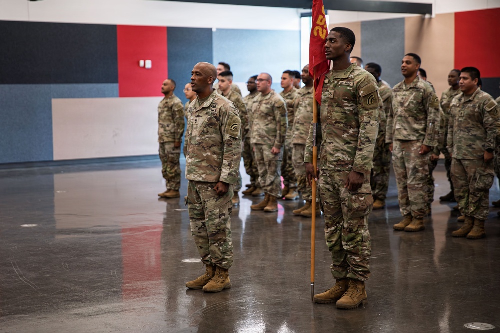 A formation of U.S. Army soldiers in Operational Camouflage Pattern (OCP) uniforms are standing at attention inside what appears to be a gymnasium or indoor facility. A soldier in the foreground is holding a unit guidon (red flag with gold lettering). The scene appears to be a formal military ceremony or formation event.