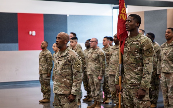 A formation of U.S. Army soldiers in Operational Camouflage Pattern (OCP) uniforms are standing at attention inside what appears to be a gymnasium or indoor facility. A soldier in the foreground is holding a unit guidon (red flag with gold lettering). The scene appears to be a formal military ceremony or formation event.