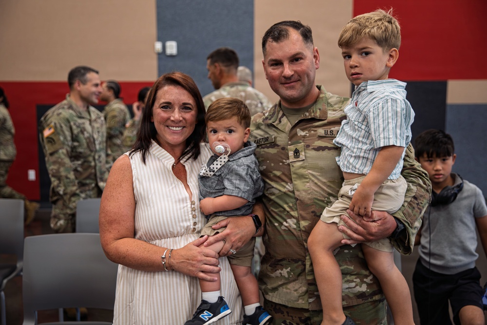 A U.S. Army soldier in OCP (Operational Camouflage Pattern) uniform poses with his family at what appears to be a military ceremony or homecoming event. The soldier, whose name tape reads 'KASTOR' and who wears a First Sergeant rank insignia, holds two young boys while standing next to a woman in a white striped dress. The background shows other soldiers and civilians in a decorated indoor venue.