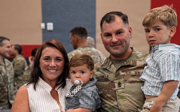 A U.S. Army soldier in OCP (Operational Camouflage Pattern) uniform poses with his family at what appears to be a military ceremony or homecoming event. The soldier, whose name tape reads 'KASTOR' and who wears a First Sergeant rank insignia, holds two young boys while standing next to a woman in a white striped dress. The background shows other soldiers and civilians in a decorated indoor venue.