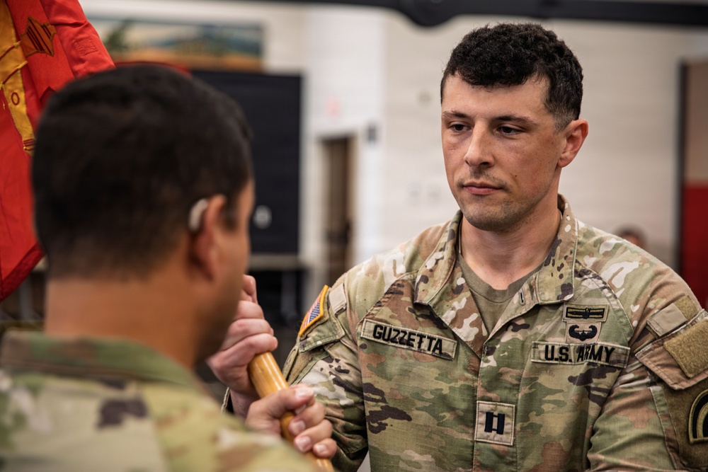 A U.S. Army Captain named Guzzetta is participating in what appears to be a change of command or guidon ceremony, receiving or passing a unit guidon staff. He is wearing Operational Camouflage Pattern (OCP) uniform with visible rank insignia (Captain's bars) and a Combat Action Badge or similar qualification badge. A red and yellow unit guidon is partially visible in the upper left of the frame.
