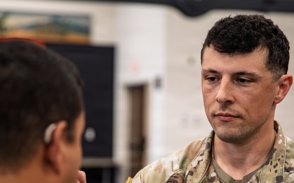 A U.S. Army Captain named Guzzetta is participating in what appears to be a change of command or guidon ceremony, receiving or passing a unit guidon staff. He is wearing Operational Camouflage Pattern (OCP) uniform with visible rank insignia (Captain's bars) and a Combat Action Badge or similar qualification badge. A red and yellow unit guidon is partially visible in the upper left of the frame.