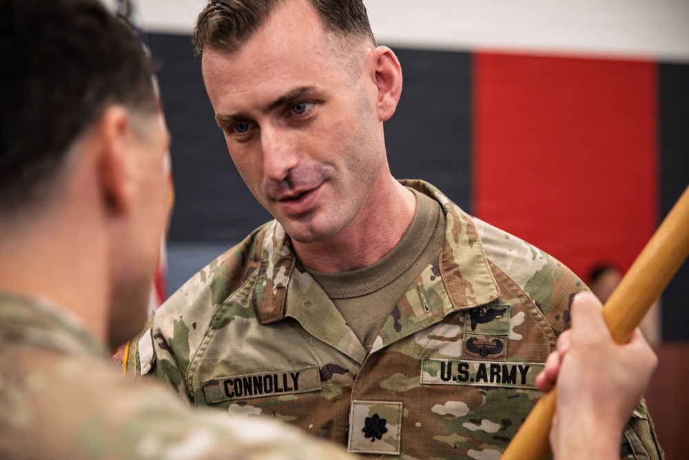 A U.S. Army soldier named Connolly, wearing Operational Camouflage Pattern (OCP) uniform, is engaged in conversation or a ceremonial exchange with another soldier whose back is to the camera. The soldier displays a Lieutenant Colonel oak leaf cluster rank insignia on his chest pocket and wears a Combat Infantryman Badge or similar qualification badge. The setting appears to be an indoor military ceremony or event.