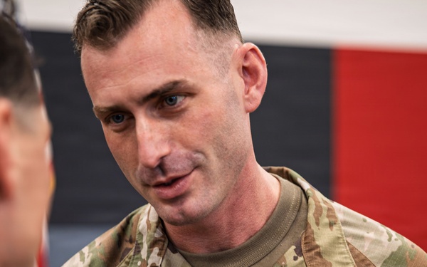 A U.S. Army soldier named Connolly, wearing Operational Camouflage Pattern (OCP) uniform, is engaged in conversation or a ceremonial exchange with another soldier whose back is to the camera. The soldier displays a Lieutenant Colonel oak leaf cluster rank insignia on his chest pocket and wears a Combat Infantryman Badge or similar qualification badge. The setting appears to be an indoor military ceremony or event.