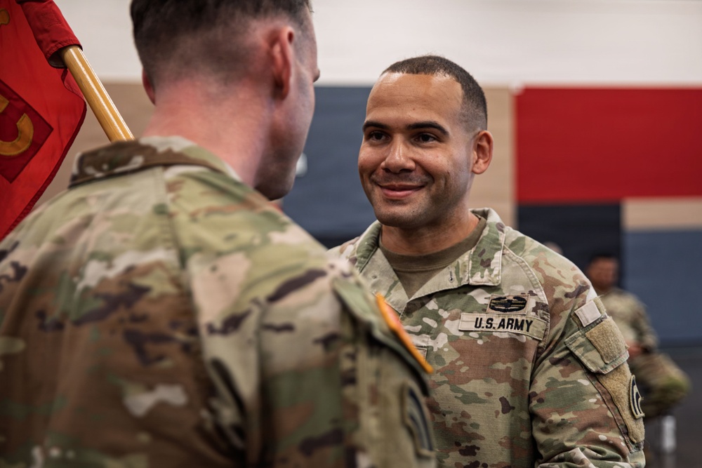 Two U.S. Army soldiers in OCP (Operational Camouflage Pattern) uniforms are engaged in what appears to be a military ceremony, likely a change of command or unit activation event. The soldier facing the camera is smiling and wearing a Combat Action Badge above his U.S. Army tape. A red guidon flag is visible in the background, indicating a formal military unit ceremony.