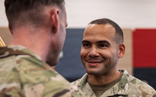 Two U.S. Army soldiers in OCP (Operational Camouflage Pattern) uniforms are engaged in what appears to be a military ceremony, likely a change of command or unit activation event. The soldier facing the camera is smiling and wearing a Combat Action Badge above his U.S. Army tape. A red guidon flag is visible in the background, indicating a formal military unit ceremony.
