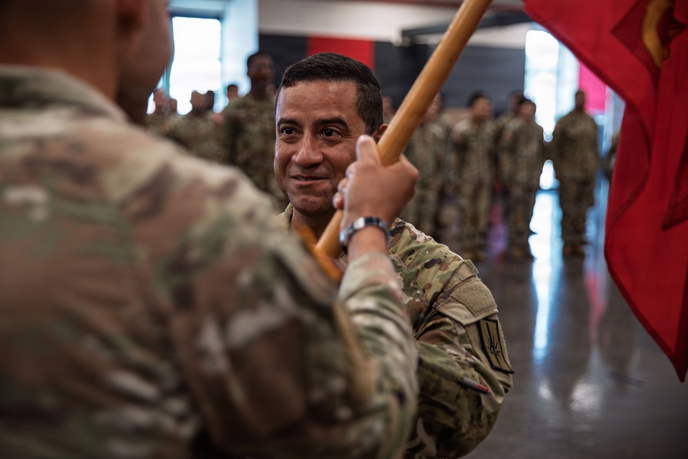 A military ceremony appears to be taking place indoors, likely a change of command or unit activation ceremony. A soldier in the foreground is receiving or passing a red unit guidon (flag) to another soldier, who is smiling. Multiple soldiers in U.S. Army OCP (Operational Camouflage Pattern) uniforms are standing in formation in the background.