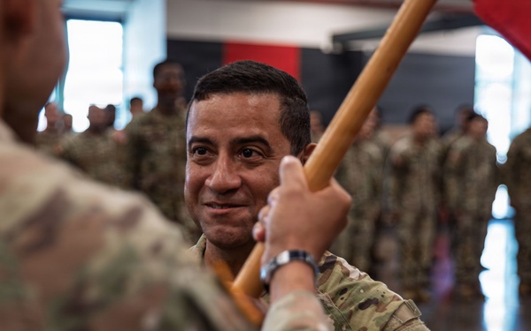 A military ceremony appears to be taking place indoors, likely a change of command or unit activation ceremony. A soldier in the foreground is receiving or passing a red unit guidon (flag) to another soldier, who is smiling. Multiple soldiers in U.S. Army OCP (Operational Camouflage Pattern) uniforms are standing in formation in the background.