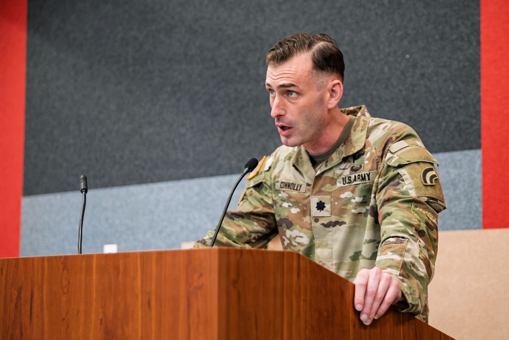 A U.S. Army Lieutenant Colonel named Connolly is speaking at a wooden podium with a microphone. He is wearing the Army Combat Uniform (ACU) in MultiCam/OCP pattern with visible name tape, branch tape, rank insignia, and a shoulder patch. The setting appears to be an indoor briefing room or auditorium with acoustic wall panels.
