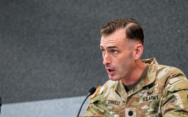 A U.S. Army Lieutenant Colonel named Connolly is speaking at a wooden podium with a microphone. He is wearing the Army Combat Uniform (ACU) in MultiCam/OCP pattern with visible name tape, branch tape, rank insignia, and a shoulder patch. The setting appears to be an indoor briefing room or auditorium with acoustic wall panels.