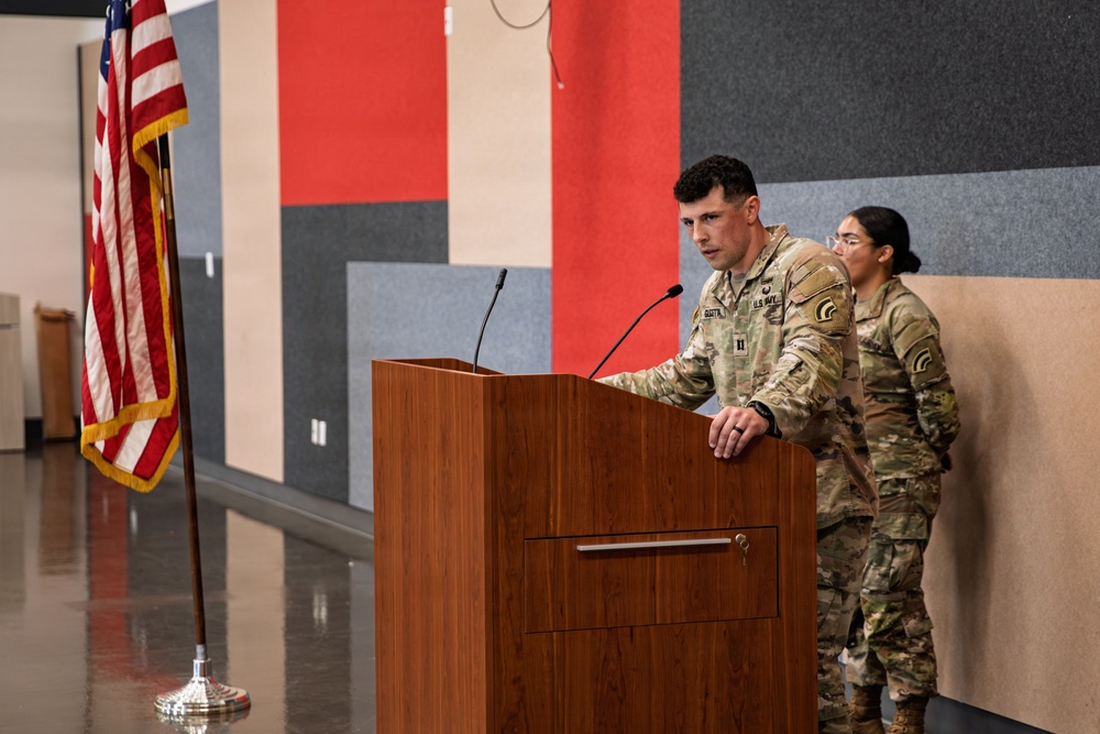 A U.S. Army Captain is speaking at a wooden podium during what appears to be a military ceremony or briefing. An American flag on a stand is visible to the left, and a second soldier stands at attention in the background. The setting appears to be an indoor facility with colorful acoustic panel walls.
