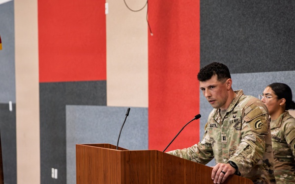 A U.S. Army Captain is speaking at a wooden podium during what appears to be a military ceremony or briefing. An American flag on a stand is visible to the left, and a second soldier stands at attention in the background. The setting appears to be an indoor facility with colorful acoustic panel walls.