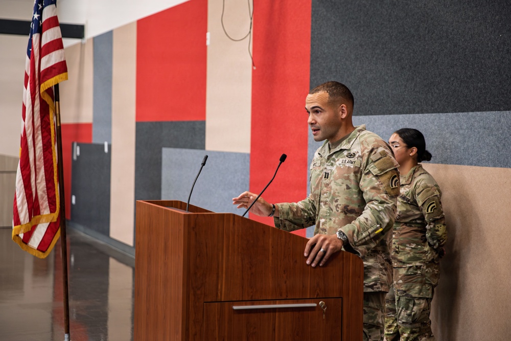 A U.S. Army officer is delivering a speech at a wooden podium equipped with two microphones. A second soldier in Army Combat Uniform (ACU) stands at attention behind and to the right of the speaker. An American flag with gold fringe is displayed to the left, suggesting a formal military ceremony or briefing.