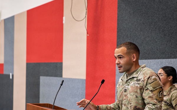 A U.S. Army officer is delivering a speech at a wooden podium equipped with two microphones. A second soldier in Army Combat Uniform (ACU) stands at attention behind and to the right of the speaker. An American flag with gold fringe is displayed to the left, suggesting a formal military ceremony or briefing.
