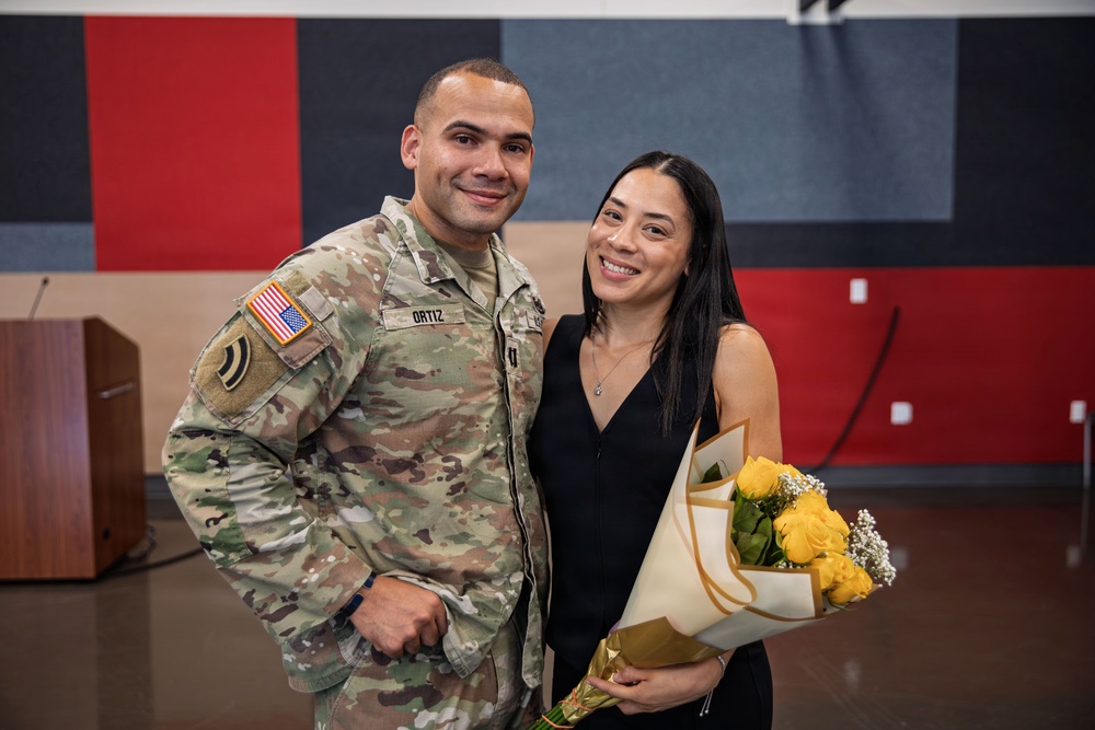 A U.S. Army soldier named Ortiz, wearing Operational Camouflage Pattern (OCP) uniform with a U.S. flag patch and unit patch, poses with a woman in a black dress holding a bouquet of yellow roses. The setting appears to be an indoor military ceremony or formal event, indicated by the acoustic panel backdrop and podium visible in the background. Both individuals are smiling, suggesting a celebratory occasion.