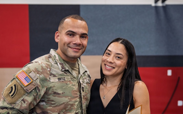 A U.S. Army soldier named Ortiz, wearing Operational Camouflage Pattern (OCP) uniform with a U.S. flag patch and unit patch, poses with a woman in a black dress holding a bouquet of yellow roses. The setting appears to be an indoor military ceremony or formal event, indicated by the acoustic panel backdrop and podium visible in the background. Both individuals are smiling, suggesting a celebratory occasion.