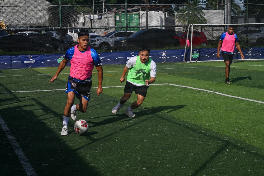 A soccer match or training session is taking place on an artificial turf field. Three players are visible, with one player in a pink bib controlling the ball while being challenged by a player in a green bib. Sponsor banners including Puma and Pacific Cup are visible along the perimeter fence.