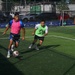 A soccer match or training session is taking place on an artificial turf field. Three players are visible, with one player in a pink bib controlling the ball while being challenged by a player in a green bib. Sponsor banners including Puma and Pacific Cup are visible along the perimeter fence.