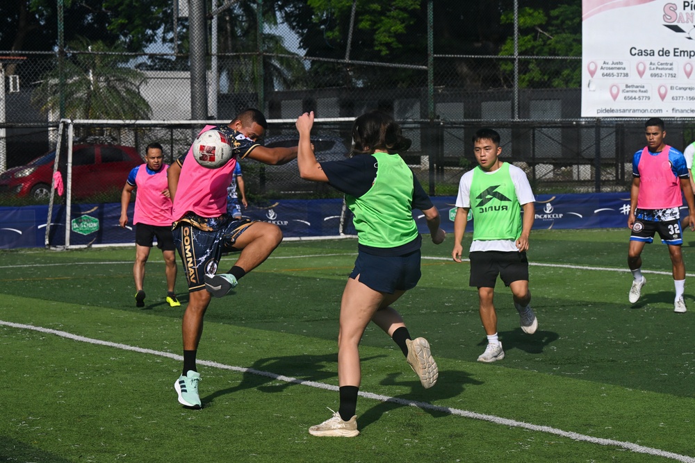 A mixed-gender recreational soccer match is being played on an artificial turf field. Two players in the foreground are competing for the ball, with one player executing a kick while the other attempts to block. Several other players and a sponsor banner for 'Casa de Empe...' (likely a financial services company) are visible in the background.