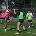 A mixed-gender recreational soccer match is being played on an artificial turf field. Two players in the foreground are competing for the ball, with one player executing a kick while the other attempts to block. Several other players and a sponsor banner for 'Casa de Empe...' (likely a financial services company) are visible in the background.