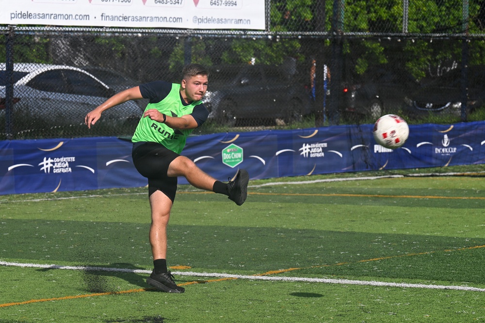A young male soccer player wearing a green vest over a black long-sleeve shirt is captured mid-kick on an artificial turf field. The ball is visible in the air to the right of the frame, suggesting it was just struck. The setting appears to be an organized event or tournament, evidenced by sponsor banners along the sideline.