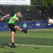 A young male soccer player wearing a green vest over a black long-sleeve shirt is captured mid-kick on an artificial turf field. The ball is visible in the air to the right of the frame, suggesting it was just struck. The setting appears to be an organized event or tournament, evidenced by sponsor banners along the sideline.