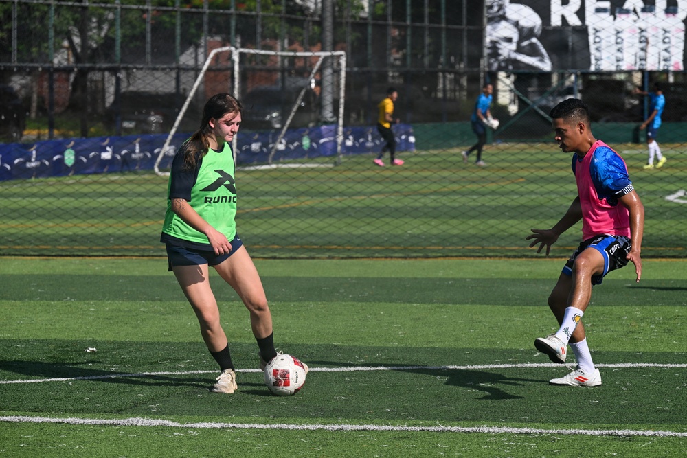 Two players are engaged in a one-on-one soccer drill or match on an artificial turf field. A female player in a green training bib controls the ball while a male player in a pink bib attempts to challenge her. Additional players and a goal are visible in the background.