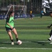 Two players are engaged in a one-on-one soccer drill or match on an artificial turf field. A female player in a green training bib controls the ball while a male player in a pink bib attempts to challenge her. Additional players and a goal are visible in the background.