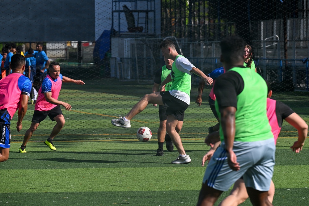 A group of players are engaged in an informal or recreational soccer/football match on an artificial turf field. Players are wearing color-coded bibs in pink and green to distinguish teams. The action is centered around a player in a green bib who is kicking the ball.