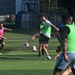 A group of players are engaged in an informal or recreational soccer/football match on an artificial turf field. Players are wearing color-coded bibs in pink and green to distinguish teams. The action is centered around a player in a green bib who is kicking the ball.