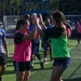 A group of people are participating in an outdoor soccer or sports activity on an artificial turf field. Two women in the foreground — one wearing a pink vest and one wearing a green vest — are giving each other a high-five in a celebratory gesture. The background shows additional players in blue jerseys, a soccer goal, and trees.