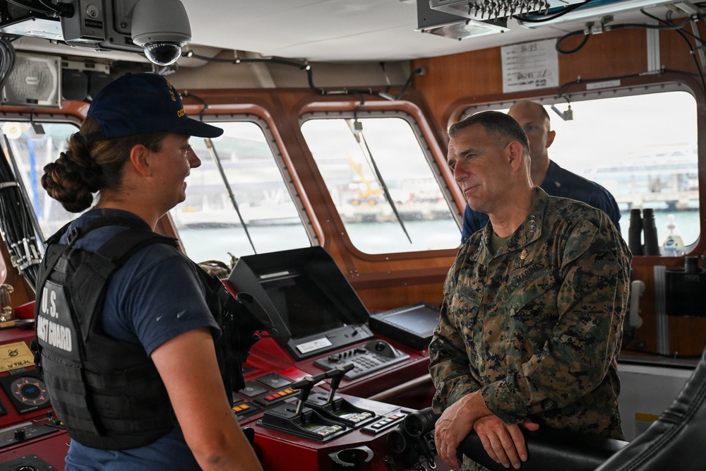 The image shows a conversation on the bridge of a U.S. Coast Guard vessel between a female Coast Guard member in tactical vest and a male U.S. Marine Corps officer in MARPAT camouflage uniform. A third military personnel member is visible in the background. The setting appears to be a ship's bridge or command center with navigation equipment visible.